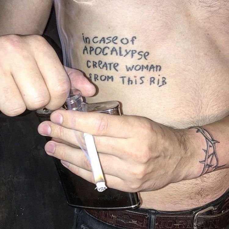 a torso with a tattoo saying “in case of apocalypse create woman from this rib.” the person’s hands are visible and are holding a cigarette and a flask.