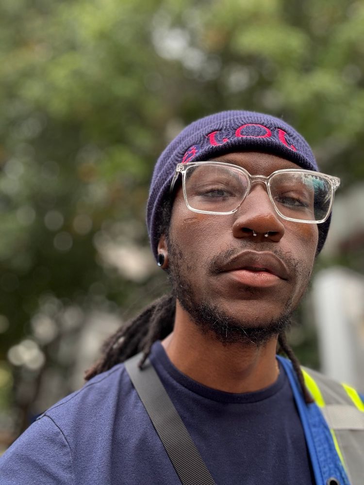 a selfie of rotelle' taken with a blurred backdrop of urbania. They're wearing clear glasses as their locs drape down their back, ever so slightly visible 