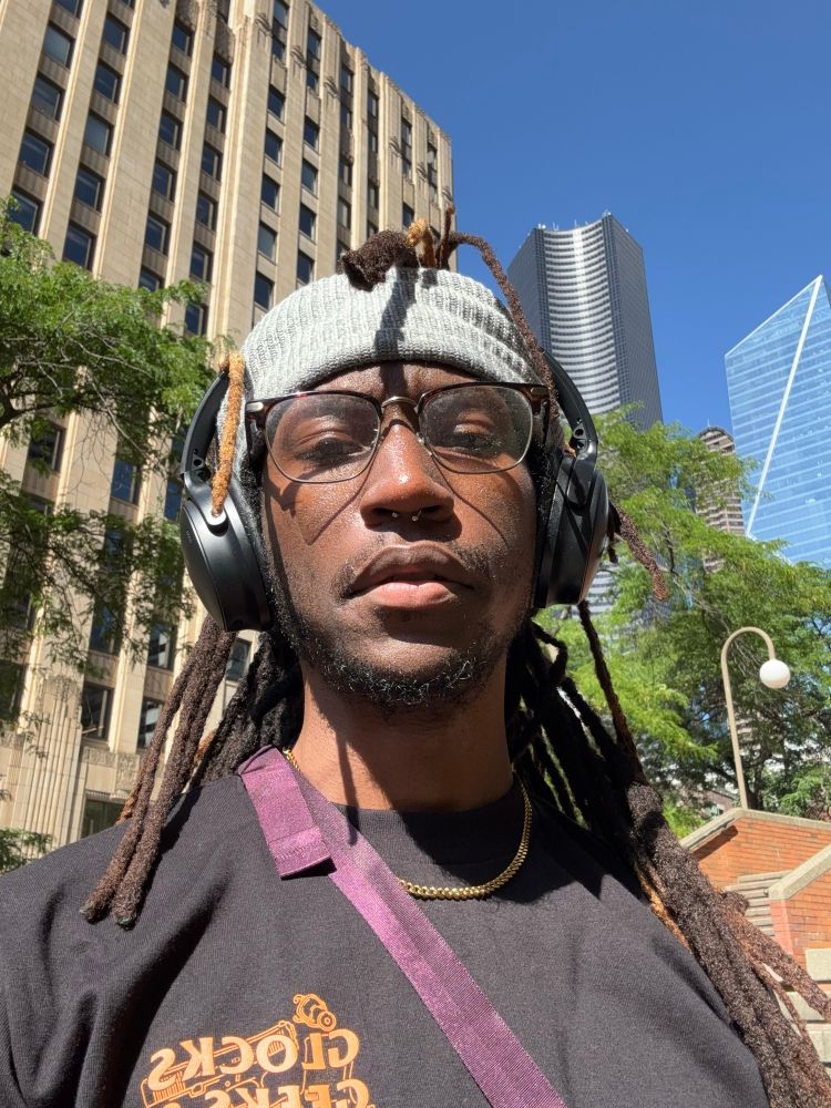Sunskissed selfie outside in the heat. its me, locs glowing as I hit a quick mean mug for the camera. My shirt is black with an orange text graphic that says "glocks, geeks, and anime"