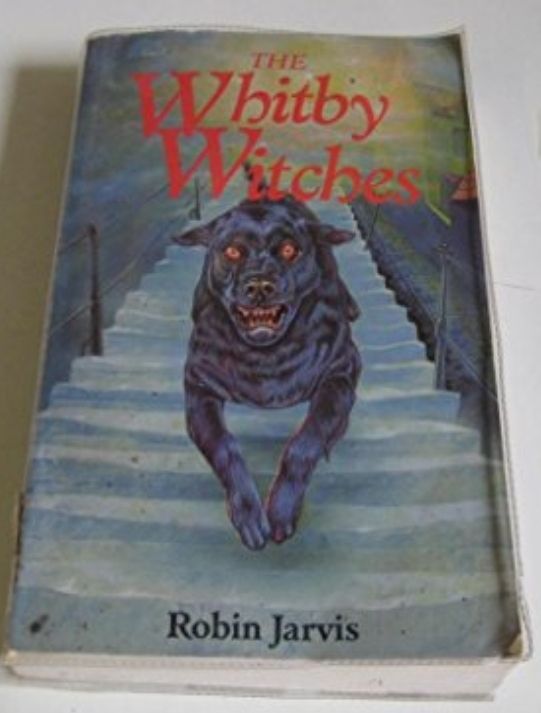 A book lies on a surface, The Whitby Witches by Robin Jarvis. The title is red text at the top, the author in black at the bottom. The illustration is of a big black dog with red eyes running down the 199 steps in Whitby. The steps are steep, old and made of stone, and therefore a little crooked and bowed, with a metal handrail up each side, and the cobbled donkey track on the right of the steps