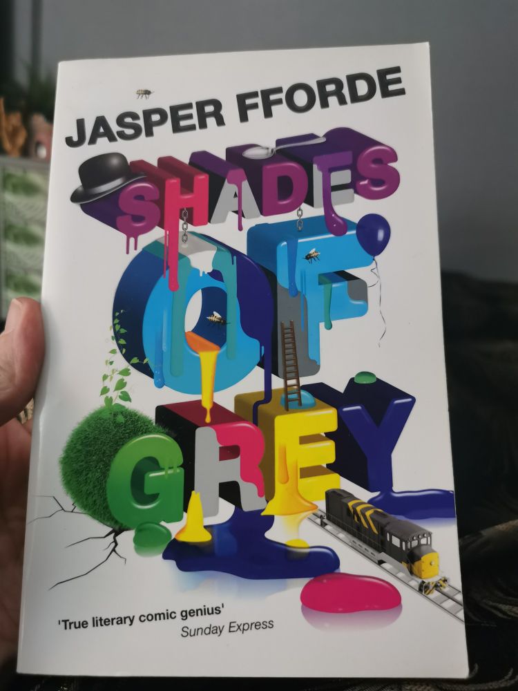 A book by Jasper Fforde titled Shades of Grey, the cover is white, the authors name is at the top in black simple text, and the title is in 3D text with each letter a different colour. There are some symbols and illustrations of a bowler hat a ladder, a balloon, paint, foliage and a train within the lettering
