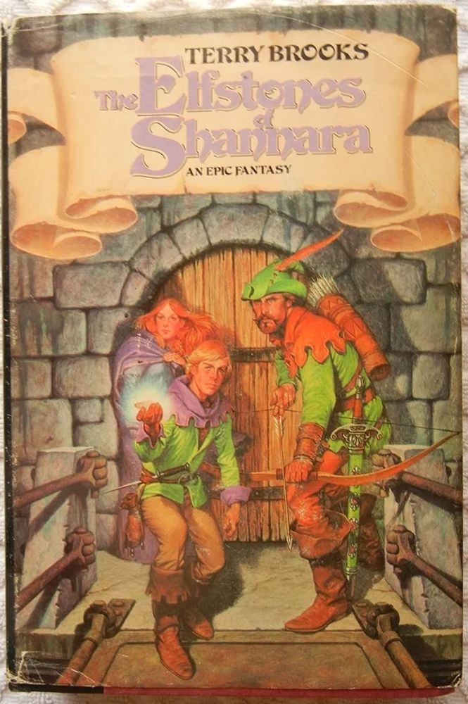 Book cover, for The Elfstones of Shannara by Terry Brooks. The text is on a scroll, and the illustration is of 3 figures in front of a castle entrance stood on a draw bridge, 1 female, 2 males, the younger male holds a pouch in his hamd and something inside is emitting a blue glow. The older male holds a bow. All are dressed in fantasy/medieval type dress