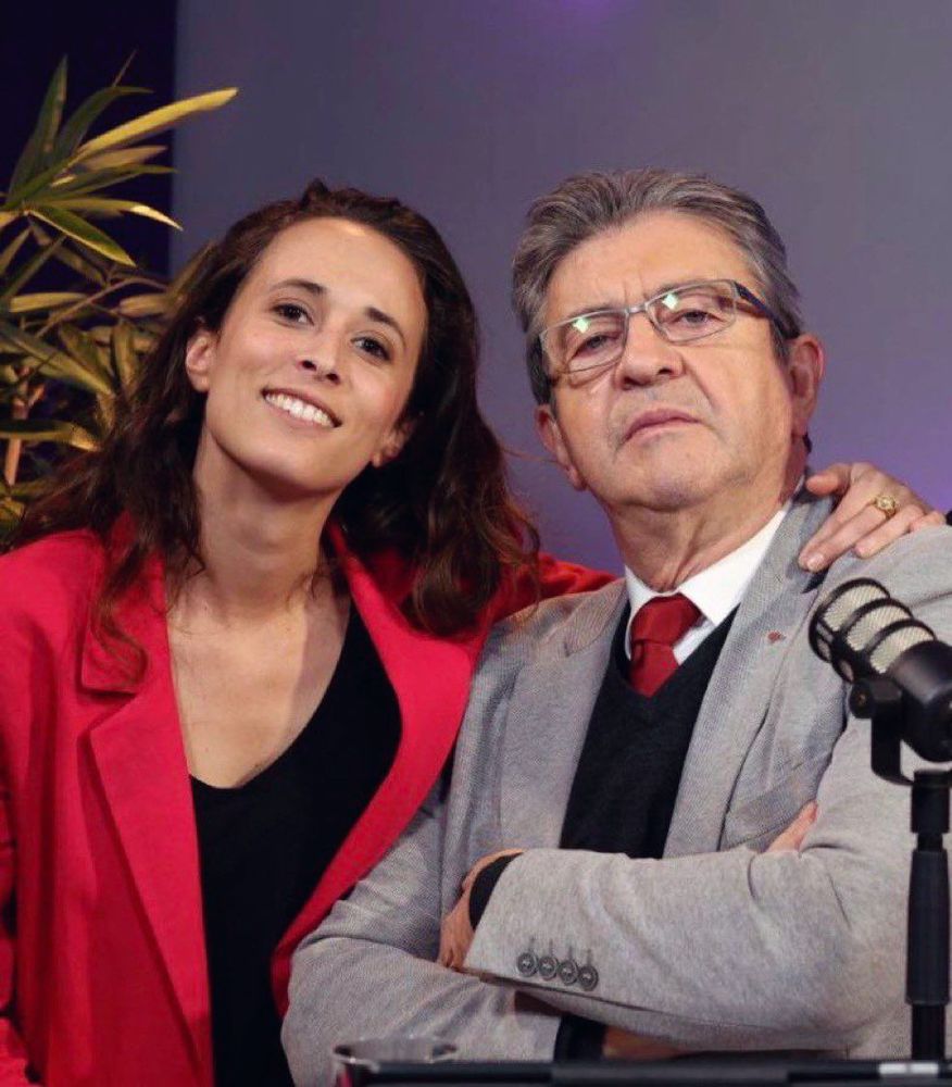 Une superbe photo de Clémence Guetté et Jean Luc Mélenchon.
Clémence est sur la gauche, elle porte une veste rouge et un t-shirt noir. Elle sourit et passe sa main autour du cou de Jean-Luc, sur la droite de l'image. Il porte un costume gris, chemise blanche, cravate rouge, pull en V noir.
Tous deux regardent droit devant eux.
Derrière eux, un fond de plateau télé, un micro sur le côté droit.