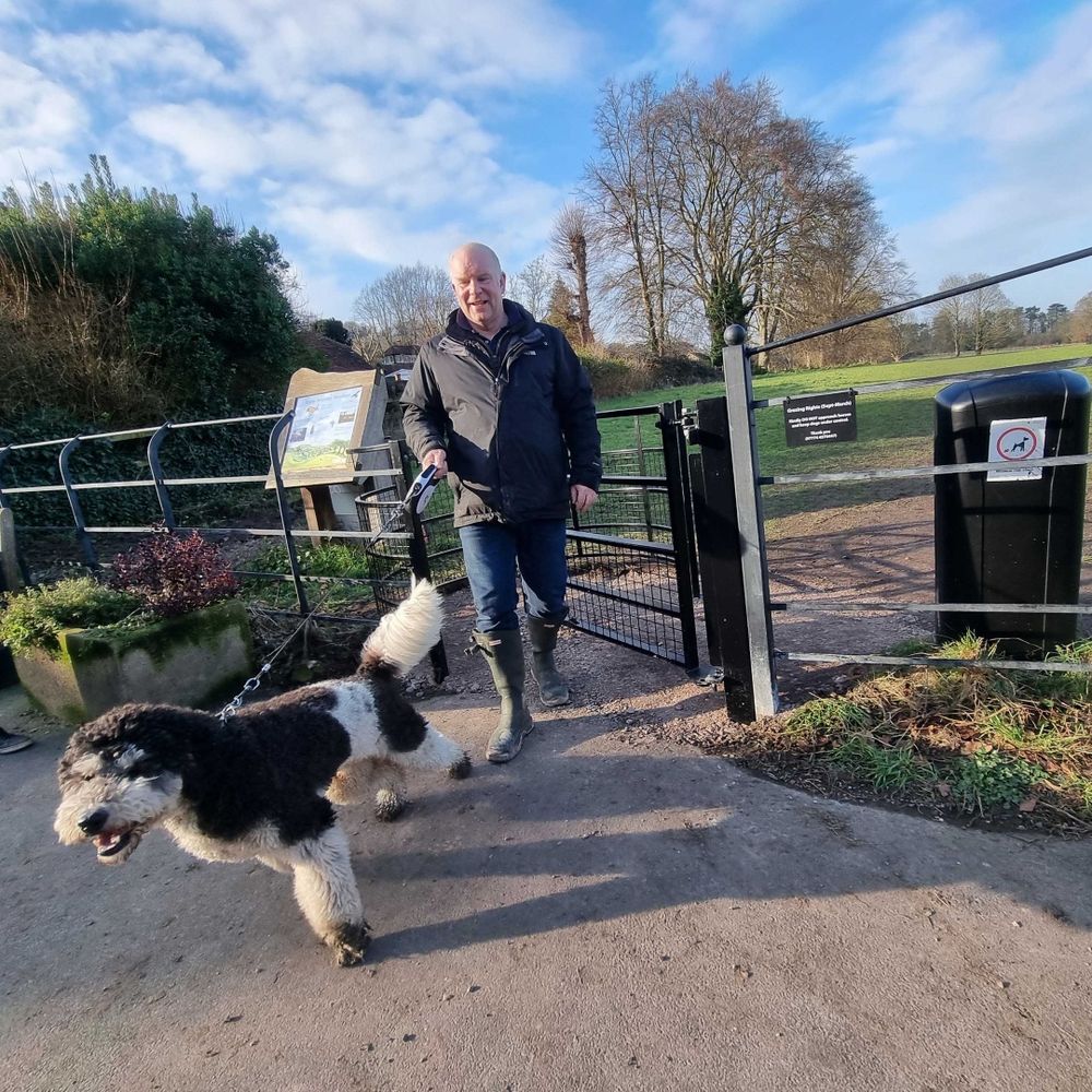 A man walking his black-and-white dog through a new gate at the entrance to a field.