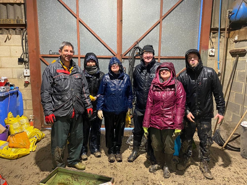 A group of 6 people stand in a barn. All are wearing waterproof coats and are soaking wet.