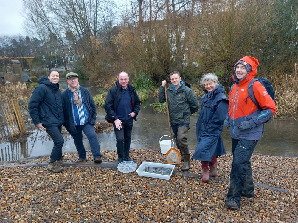 A group of 6 people stand on a pebbled river bank.