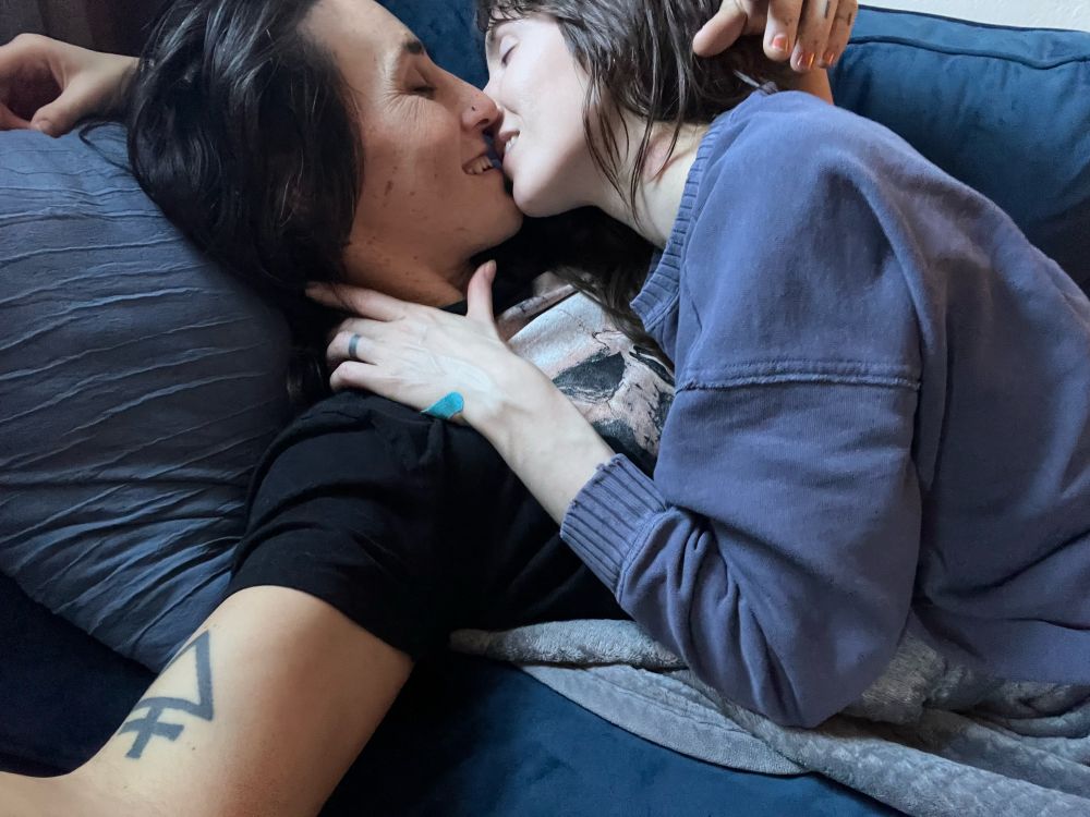 On a blue velvet sofa bathed in sunlight a queer couple lie sprawled, smiling into a kiss. 