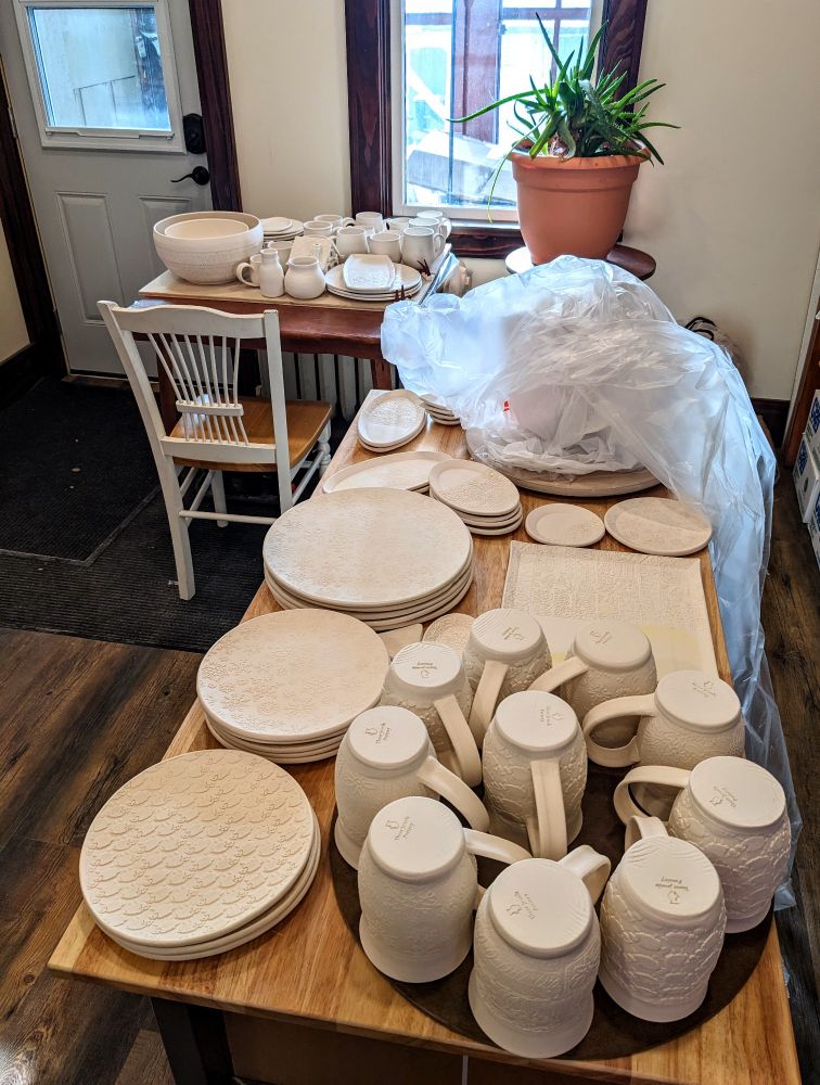 Stacks of white, unglazed ceramic pottery sit on two tables (mugs, plates, bowls, etc.). There is a large aloe plant in a terracotta-colored pot that is spiking out in several directions. 