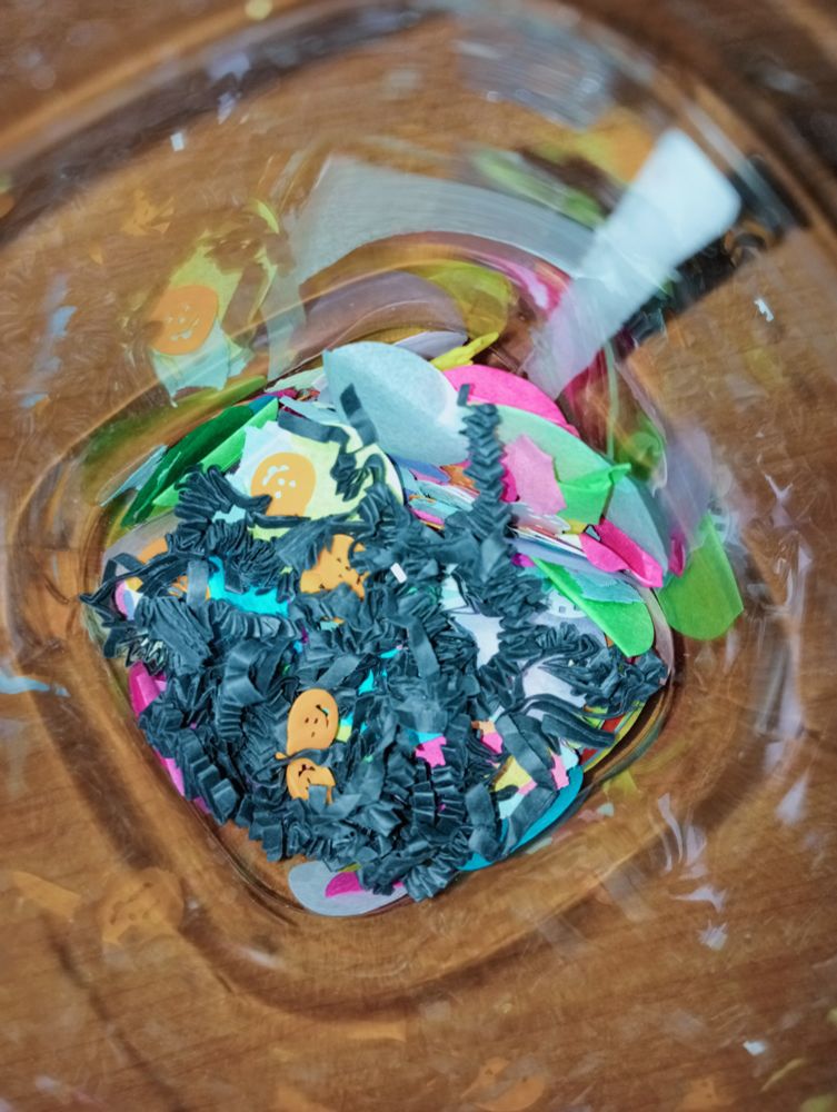 Interior de un bote de plástico transparente en cuyo fondo hay confeti. Se ve en la parte de arriba del todo tiras de papel negro arrugadas y calabazas de halloween.