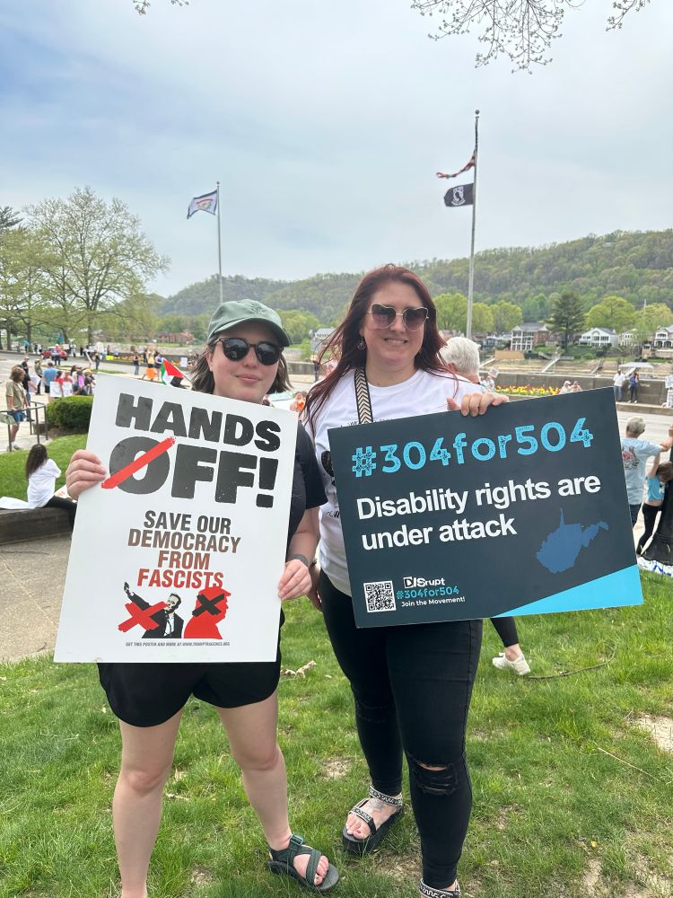 Two women standing on a grassy area. One woman on the left is wearing a gray cap, sunglasses, black T-shirt, and shorts, holding a sign that reads, "HANDS OFF! SAVE OUR DEMOCRACY FROM FASCISTS" with silhouettes of people marked with red Xs. The woman on the right is wearing sunglasses and a white T-shirt, holding a sign that says, "#304for504 Disability rights are under attack" with the shape of West Virginia depicted. Behind them, a crowd is visible with flags in the background.