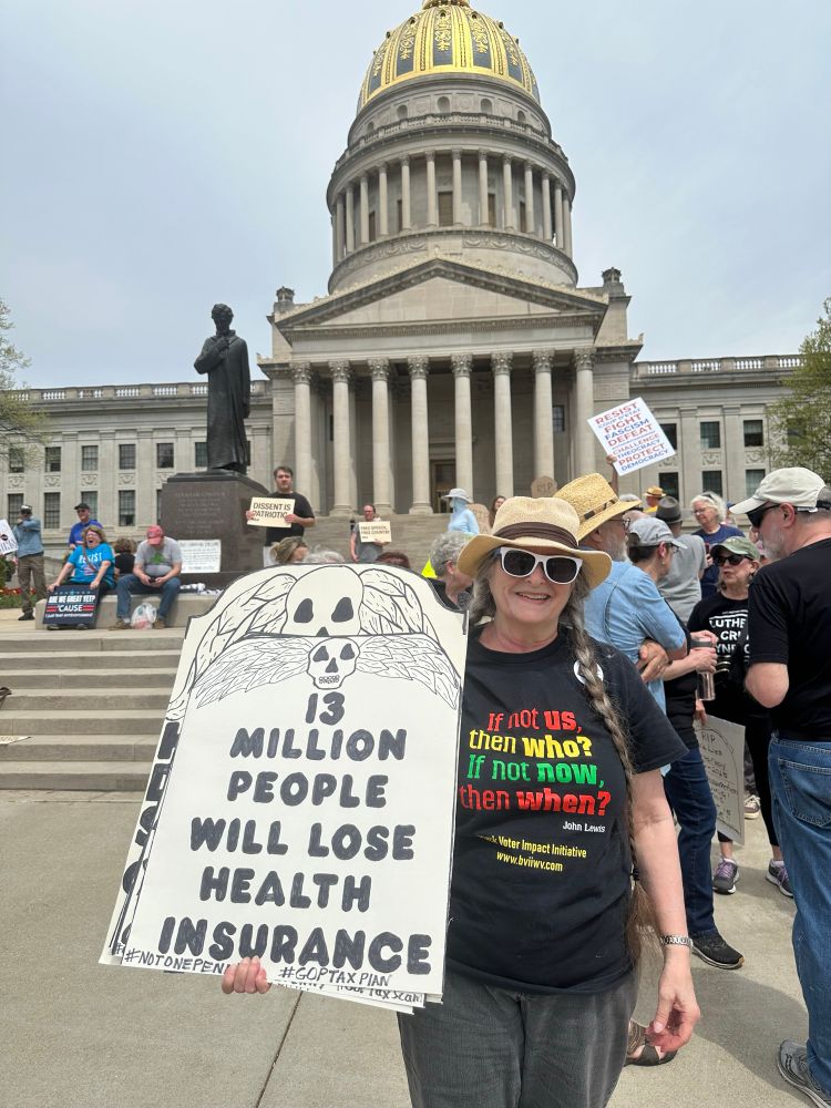 A woman is holding a large sign while standing on the paved area in front of the building. The sign features a drawing of a skull and wings, with bold text “13 million people will lose health insurance,” underneath. The woman is wearing a sun hat, sunglasses, and a black t-shirt featuring a colorful quote. Behind her, a group of people is gathered, some holding placards with various messages.