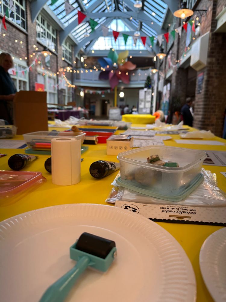 A table of craft materials set up in Weston Museum underneath the glass ceiling. The table is yellow, and in the foreground are printing inks, and an ink roller on a white paper plate. The background is out of focus but contains coloured papers, pencils and in the distance are stings of fairy lights and bunting. 