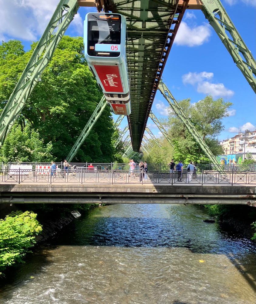 Wuppertaler Schwebebahn: Foto von unterhalb der Bahn. In der Mitte der Fluss mit einer Brücke, darüber die Metallkonstruktion der Bahn und ein Zug. 