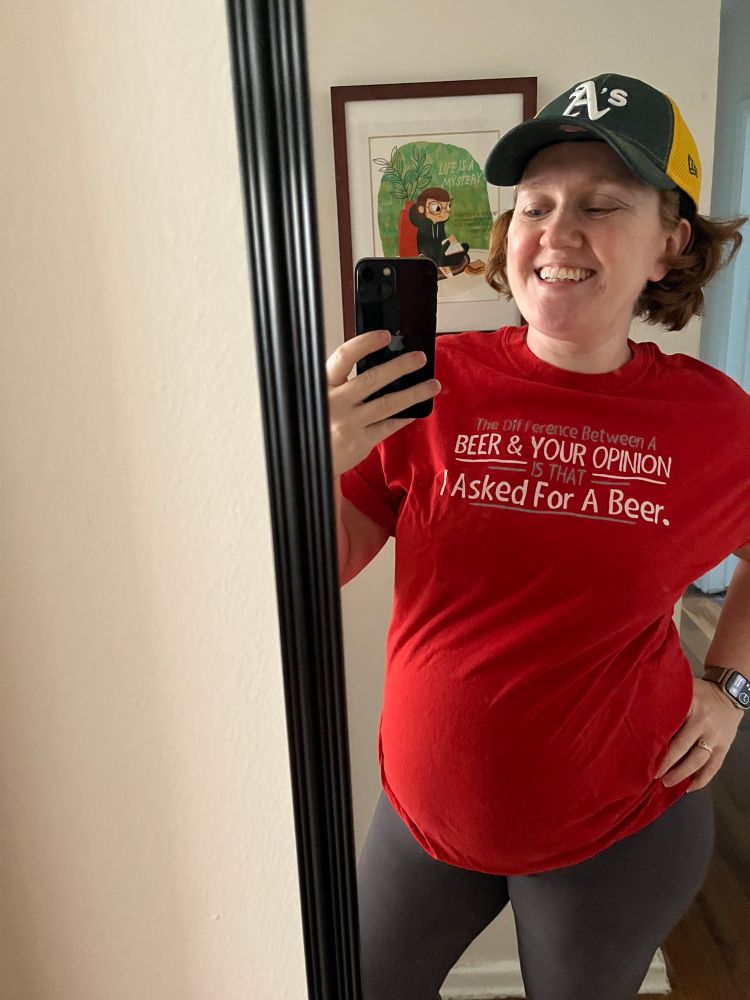 A pregnant woman wears a red shirt that says, “The difference between a beer & your opinion is that I asked for a beer.”