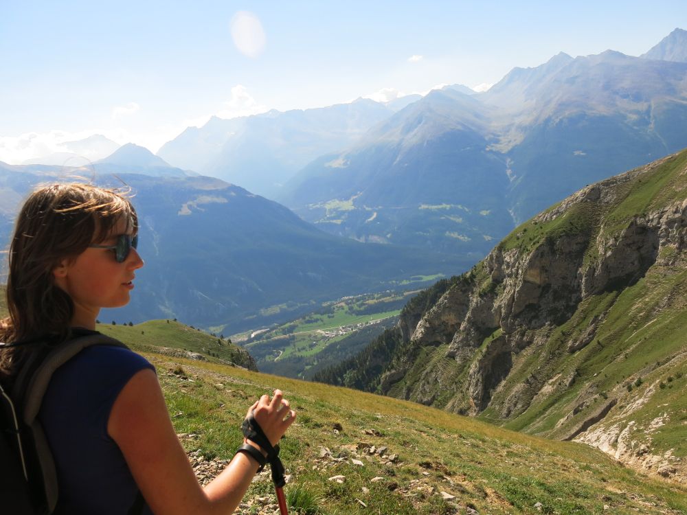 Roxanne Armfield looking out on the french Alps in the summer. Roxanne is in the foreground with grass-covered hilltops behind her. Further back are dark green mountains lightly covered by clouds. 