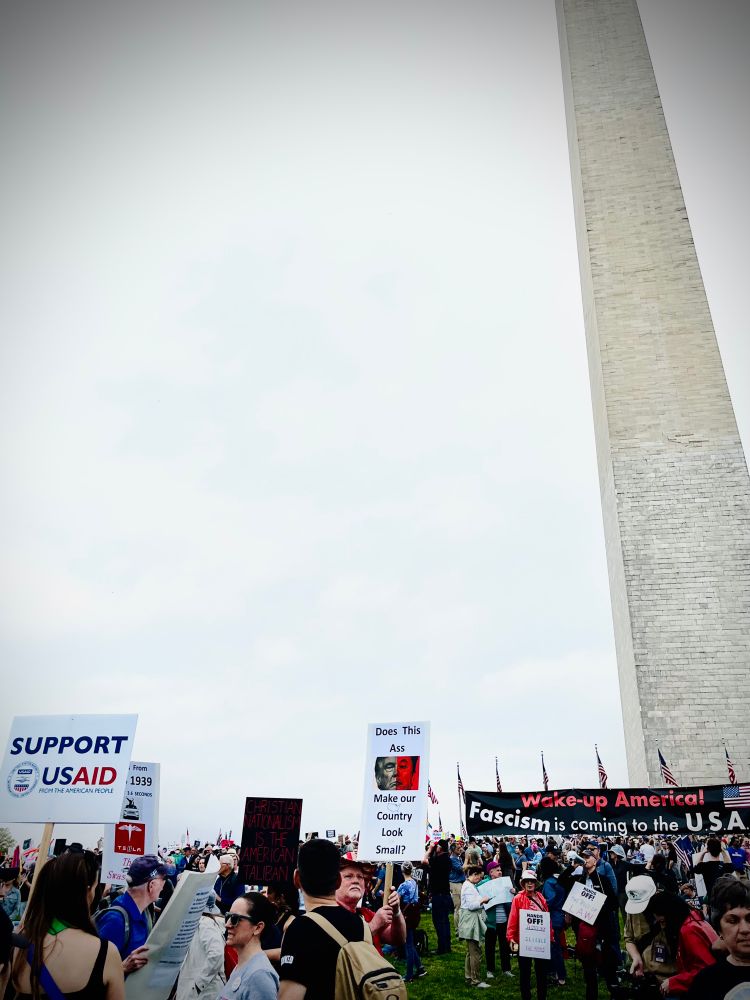 Protesters in front of the Washington monument. Protest signs read: “Support USAID” and “Wake up America! Fascism is coming to the USA”.
