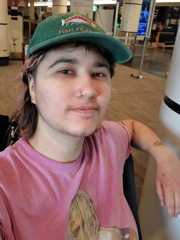 Selfie of an androgynous person with a green cap and a pink shirt featuring Britney Spears.