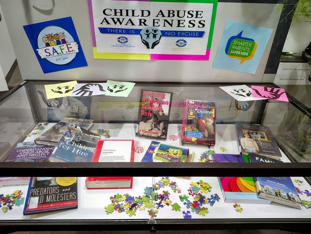 The table-top display case has a glass top and sides with 10 books, a puzzle box, and puzzle pieces strewn about inside. On the wall behind and above the display case, there are three signs, and the center one is from the Safe Harbor Children's Advocacy Center and says, "Child Abuse Awareness: There is no excuse." There are 5 images on top of the display case portraying open or holding hands on square, colored paper.