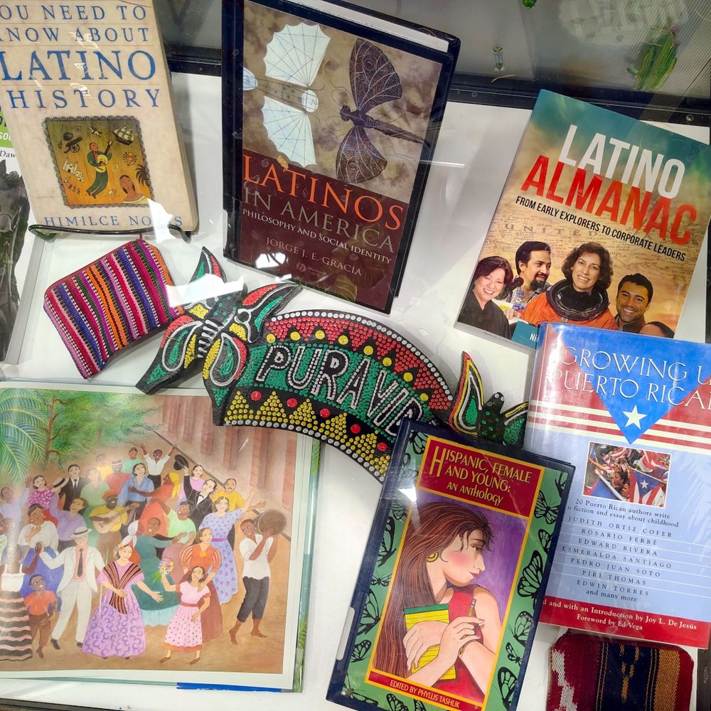 Books on the right half of the display case are Everything You Need to Know about Latino History; Latinos in America: Philosophy and Social Identity; Latino Almanac: From Early Explorers to Corporate Leaders; Hispanic, Female, and Young: An Anthology; and Growing Up Puerto Rican. There is also a banner that says, "Pura Vida." 
