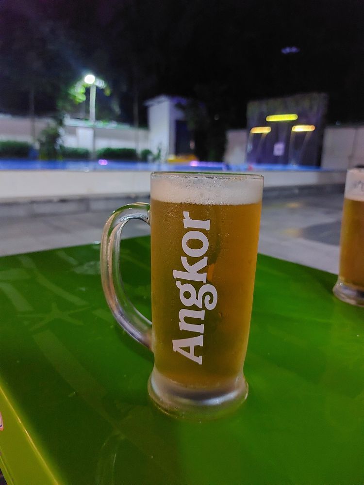 Een glas bier, op het glas staat Angor.
Het glas staat op een groen tafeltje. 
Op de achtergrond is vaag het zwembad te zien.