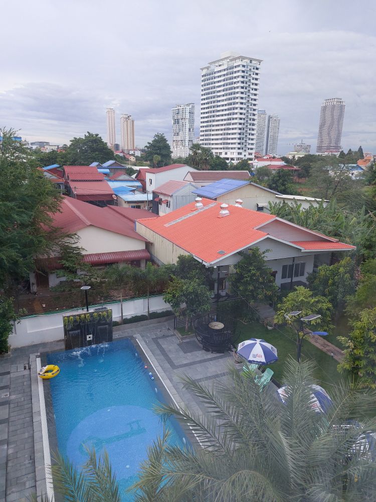 Uitzicht vanaf de bovenste verdieping van het hostel. 
Beneden is een zwembad met palmbomen, parasols en stoelen.
Daarachter huizen en daarachter hoge toren flats.