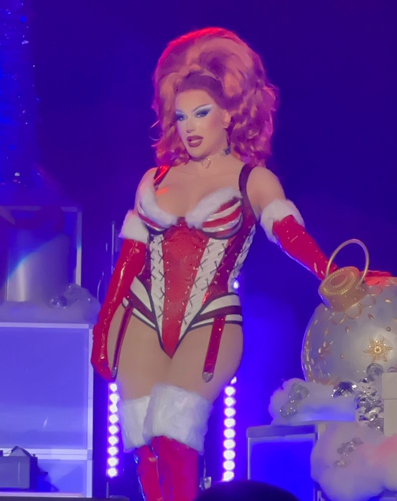 Plane Jane in a red and white corset outfit with long red gloves that almost go to her shoulders. Her boots match the gloves. She has on a red wig with a high pony