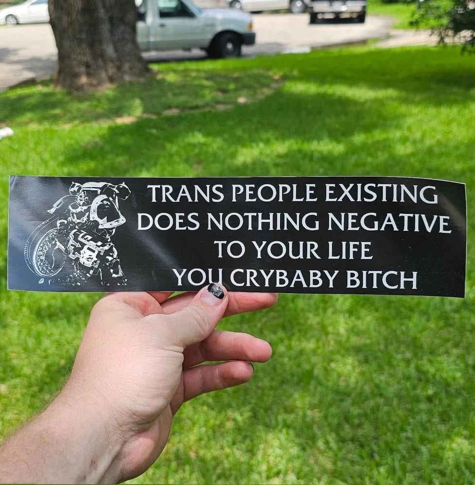 Black bumper sticker that says “TRANS PEOPLE EXISTING DOES NOTHING NEGATIVE TO YOUR LIFE YOU CRYBABY BITCH”