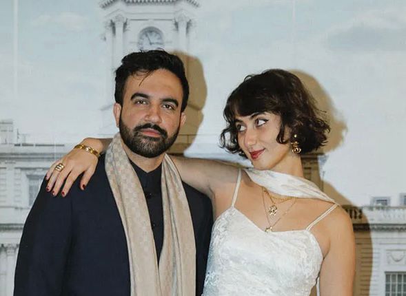 mamdani not smiling standing next to his wife, standing, who is absolutely beaming up at mamdani 