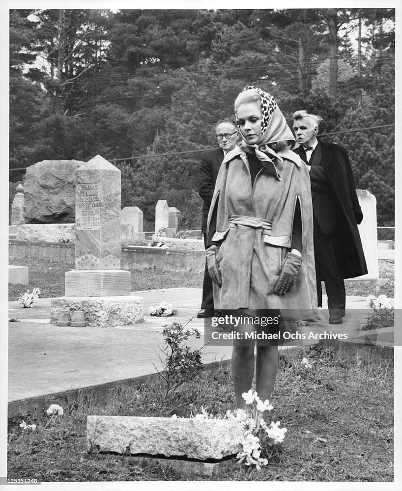 Sandra Dee in a cemetery wearing what appears to be a faux leather coat without sleeves. It’s got slits on either side to shove her arms out. The shape of the coat could be compared unfavorably to that of a garbage bag.