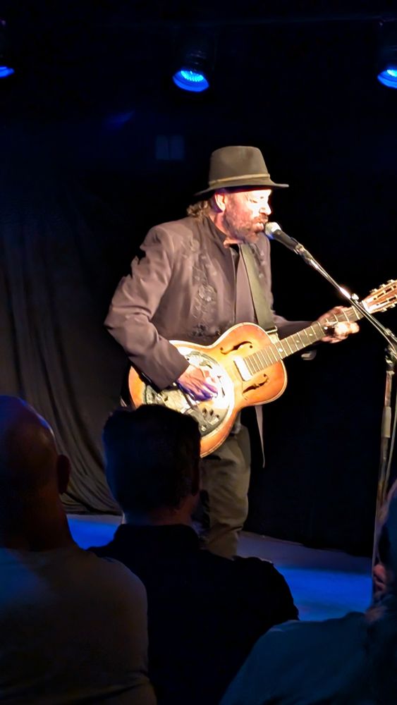 Blues musician Colin Linden playing solo on stage in a basement club. He's dressed in black and playing a steel guitar, well lit by stage lighting. 