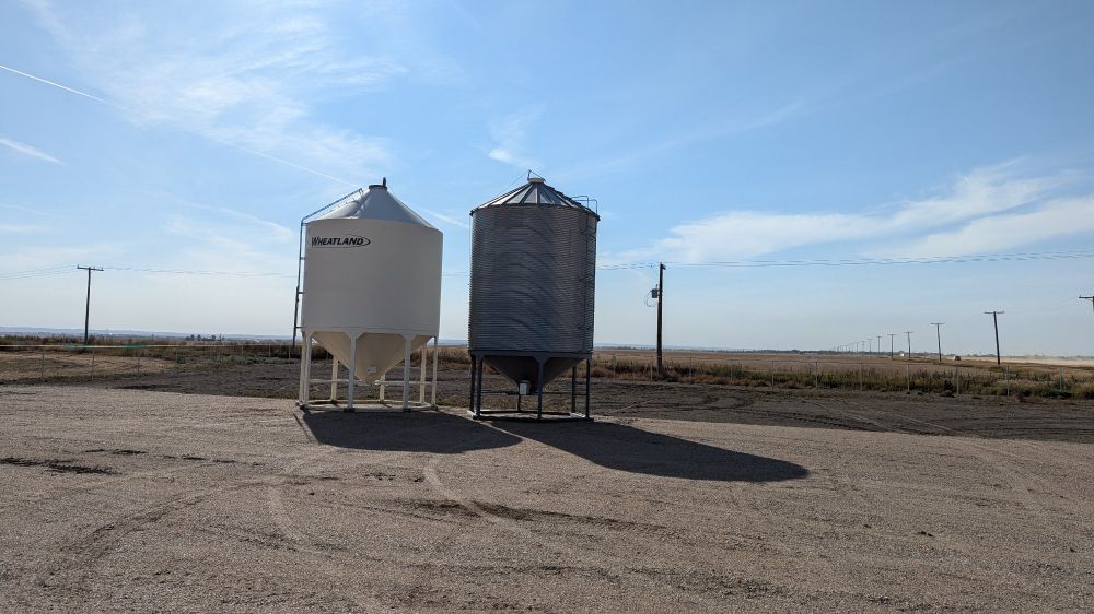 Two hopper bottom grain bins in the middle of some open yard space at the SaskPolytech Agriculture and Food Production demonstration farm SW of Moose Jaw. Not a wind blocking tree to be found for hundreds of km to the west.