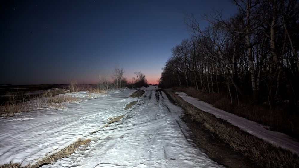 Dead end with a road covered in snow and mud. It is now late evening and the sky is dark purple with a line of fading orange along the eastern horizon. There is a line of trees on the north (right) side of the dirt road. 