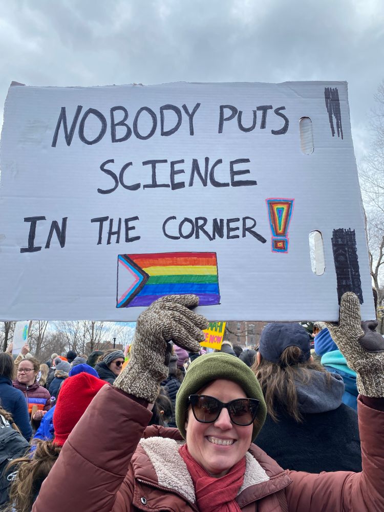 Woman at the Stand Up for Science rally in Boston with sign that says “Nobody puts science in the corner!” And the Pride flag on it. 