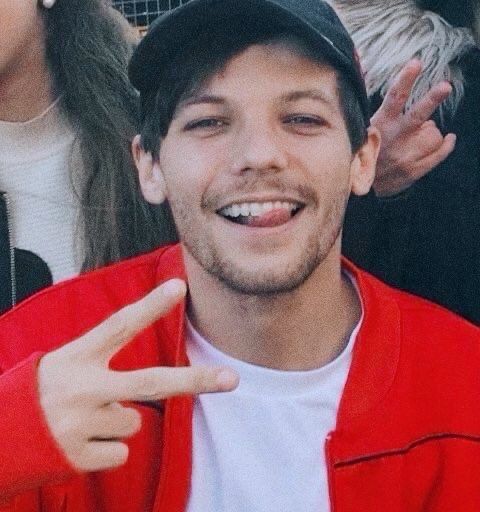 Louis wears a black cap and a festive bright red jacket over a white t-shirt. He has his tongue sticking through his teeth, tongue pointed toward the front and a bit off to the side, and his hand is making the V sign or backwards peace sign. He is using his right hand and there are no visible tattoos.