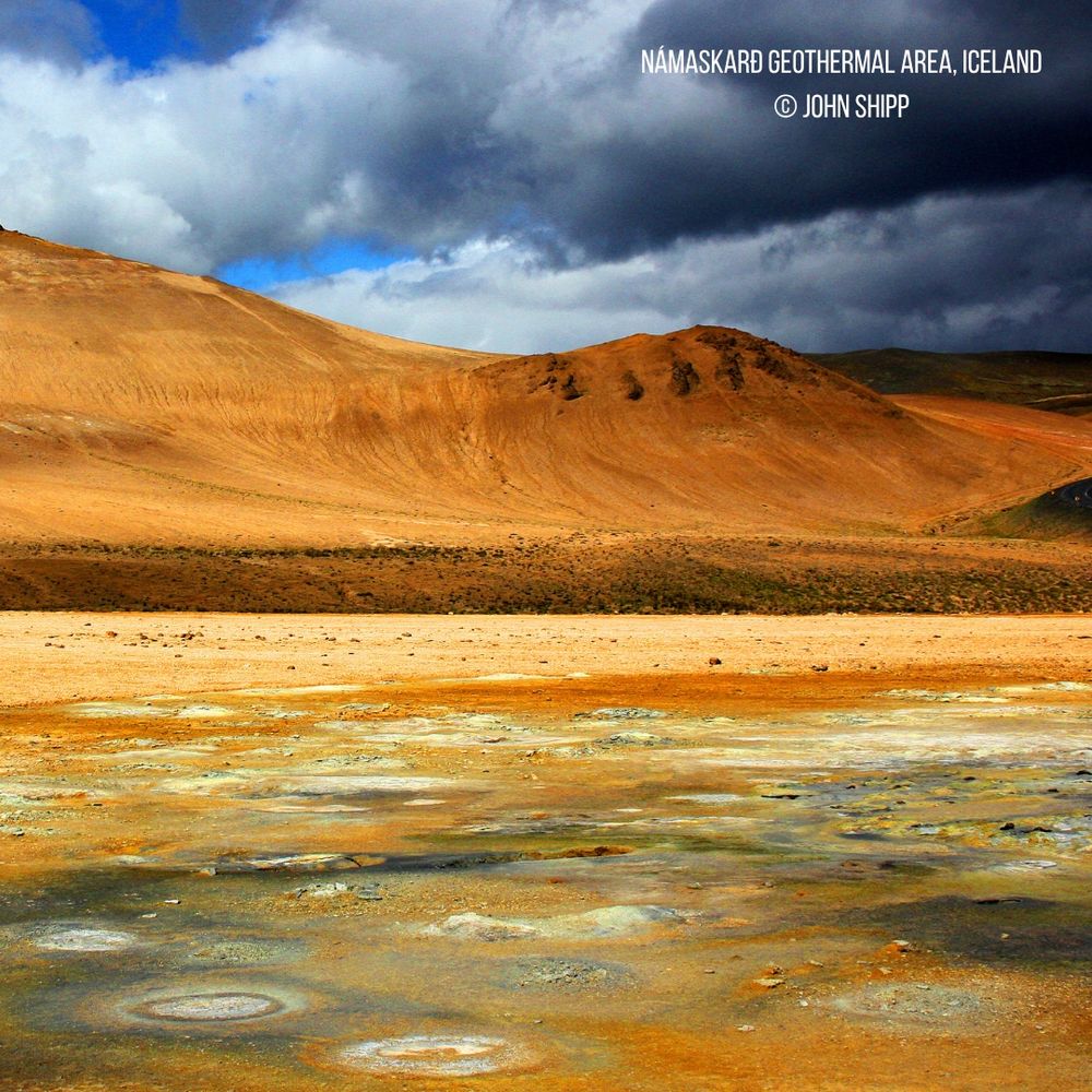 Namaskard Geothermal Area, Iceland. Namaskard is a colourful area of hot springs and boiling mud pools near Myvatn in north-east Iceland. In 1969 a geothermal power plant was build to the west which was one of the first geothermal stations in the world to produce electricity. © John Shipp. In the foreground is a colourful, flat geothermal plane in shades of terracotta, white and grey with occasional concentric circles where the mud pools have formed. In the distance is a terracotta-coloured mountainside devoid of vegetation sitting below a sky with dark, threatening clouds.