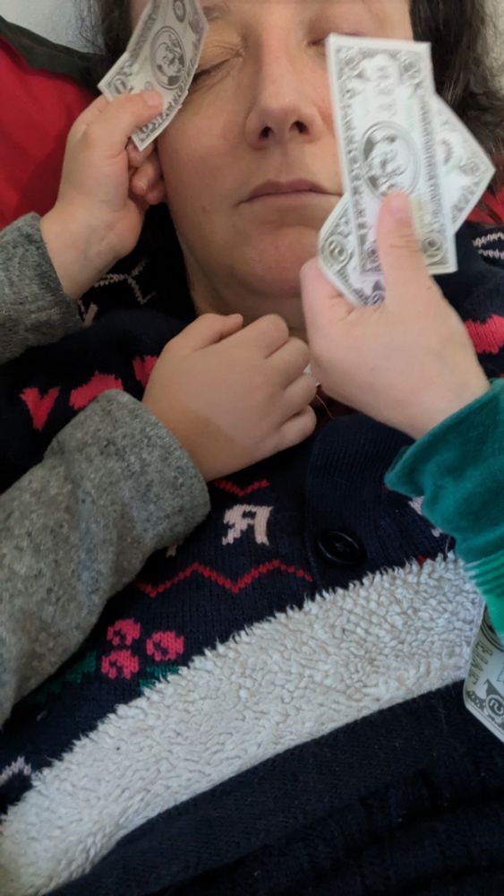 Eu mulher branca de cabelo moreno de olhos fechados e dois pares de mãozinhas de crianças com notas de brinquedos sendo esfregadas no meu rosto 