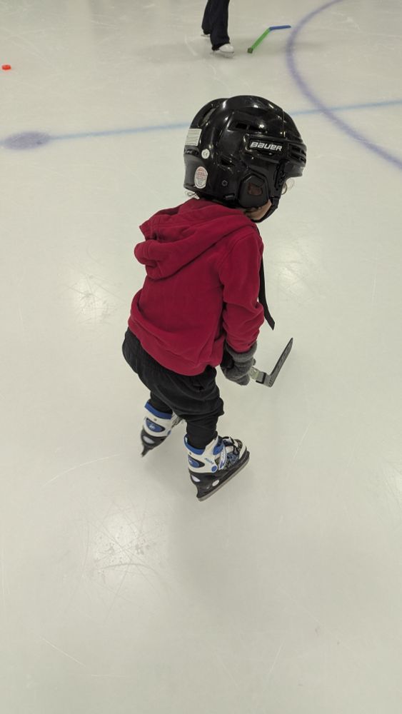 Criancinha no gelo de blusa vermelha calça preta e capacete preto segurando um taco de hockey 