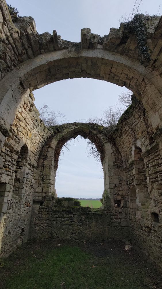 Une nef d'église en ruine. L'espace pour le vitrail est vide et permet de voir le ciel et un champ en arrière-plan. Le toit a lui aussi disparu et une faible végétation pousse sur le haut des pierres. Au sol, la terre est recouverte de plantes et crée un tapis vert.