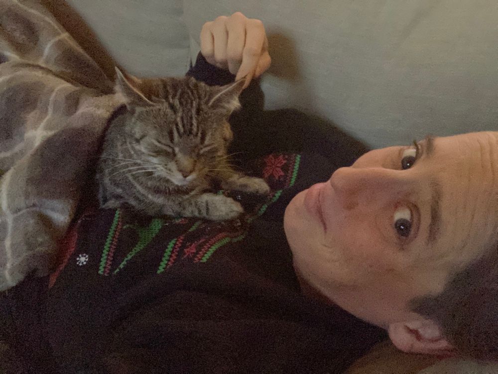 A pale skinned androgynous person in a black sweatshirt is lying on their back on a couch with a small tabby cat sleeping on their chest. A blanket is loosely draped over both cat and their chest. 