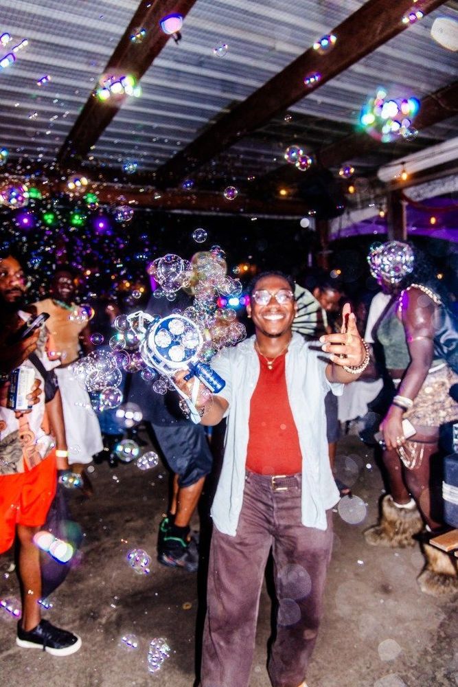 Seven, a DJ, smiles and laughs while pointing at the camera. He holds a bubble gun.
