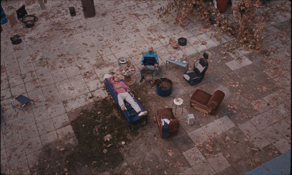 Two men recline on chairs outside. They are surrounded by fallen leaves.