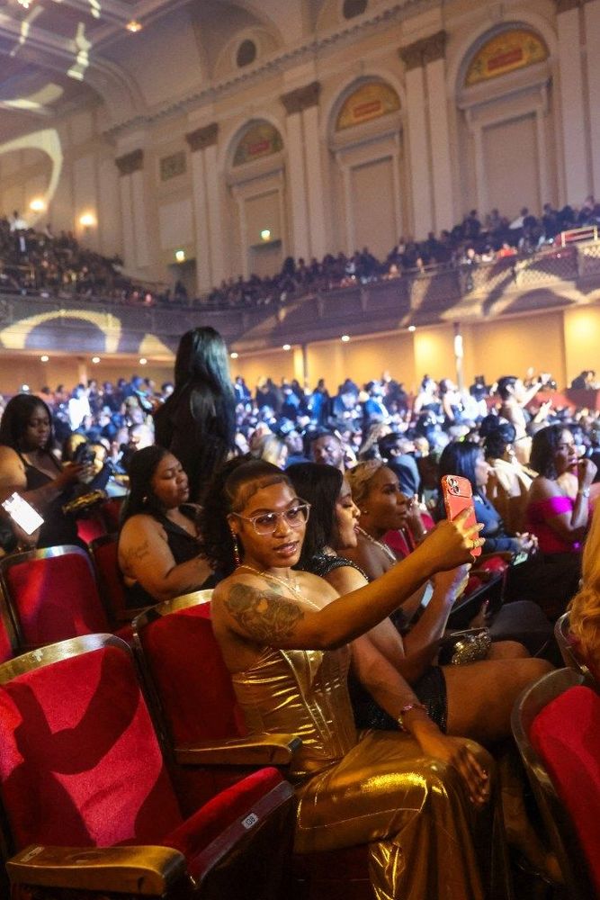 A woman in a gold dress sits in the audience. She has on a gold dress and is holding up her cell phone.
