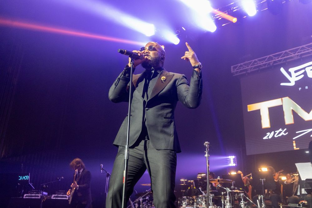 A Black man dressed in an all black suit and wearing sunglassess performs on stage.
