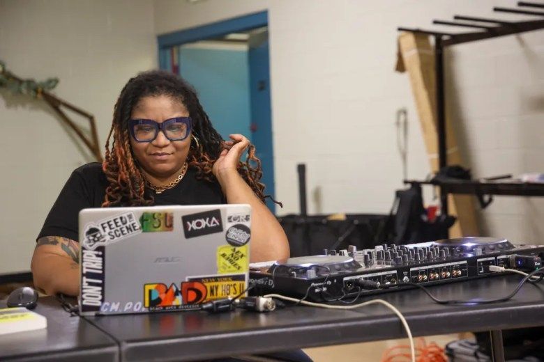 A dj with brown skin and locs is at a computer and her dj set up