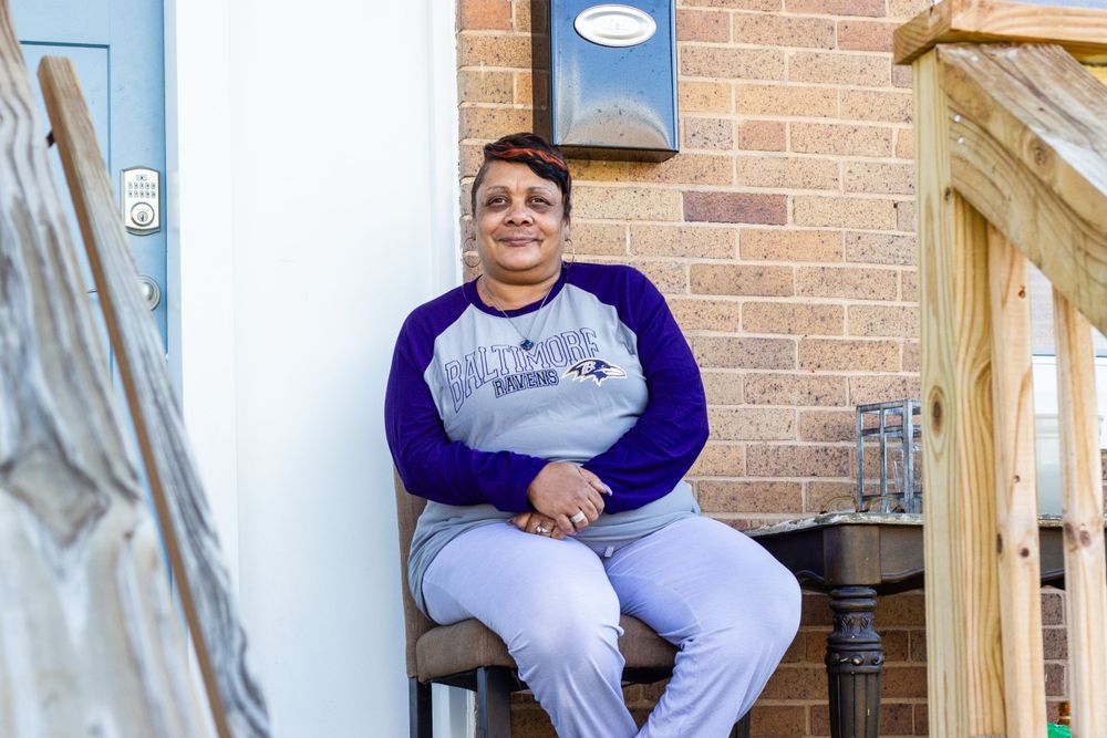 A Black woman with short hair poses for a photo. She is wearing a purple, gray, and white Baltimore Ravens shirt and light colored pants.