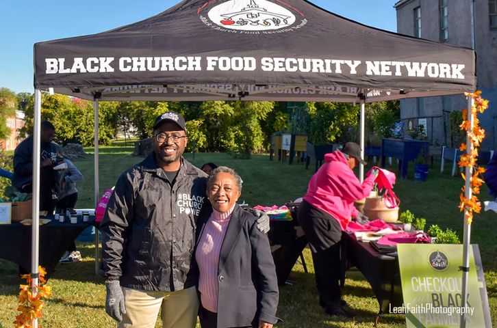 Two people, a man and a woman, stand together under a black tent. Words on the tent read: "Black church food security network."