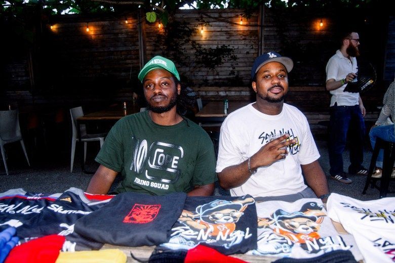 Two men sit at a table. A variety of t-shirts for sale are folded on the table.