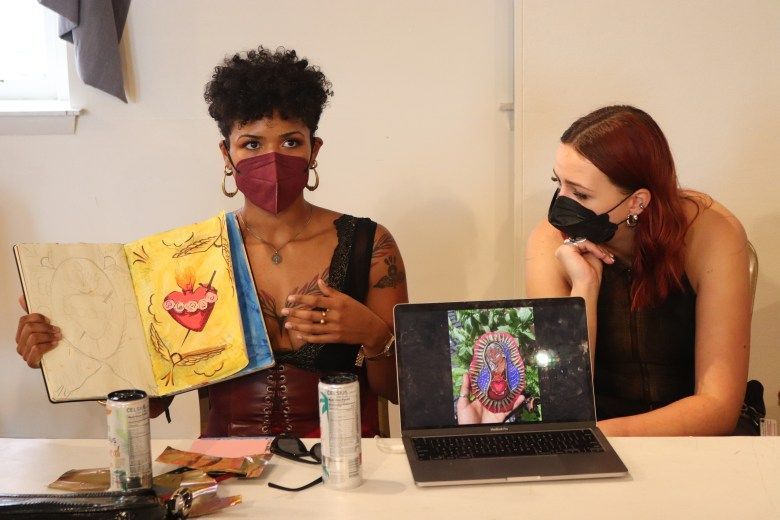 Two people. One, wearing a face mask, holds an open book. The other looks on.