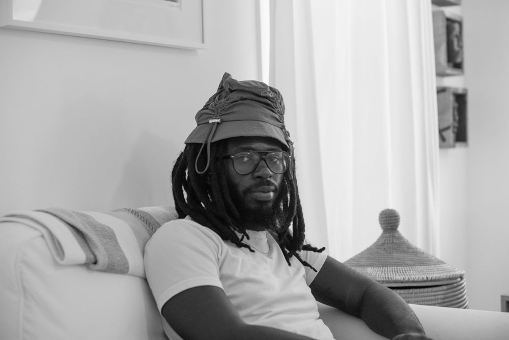 a person with brown skin sits in a living room, he is wearing a hat