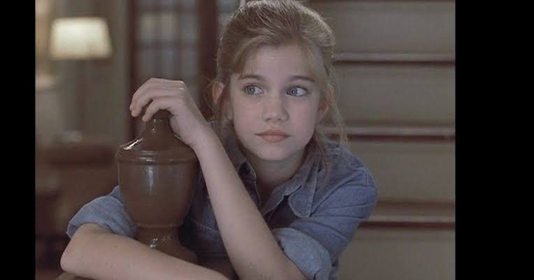 Frame de filme (acho que Meu Primeiro Amor). A atriz loirinha, com o cabelo amarrado com fios soltos, está sentada num degrau de escada, segurando no corrimão e olhando para o lado, vestida com uma camisa de botão jeans.